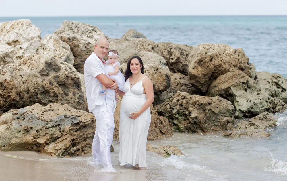 A family in white on the beach rocks, mom expecting, dad holding their baby girl